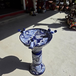 May include: A ceramic birdbath featuring a deep blue and white floral pattern. The basin has three small sculpted birds perched on the rim. The pedestal is cylindrical and flares at the base, with matching intricate blue and white floral designs.