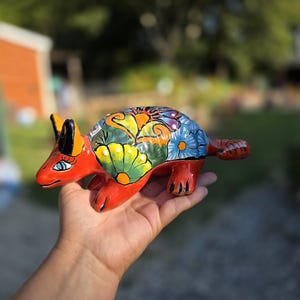 May include: A ceramic armadillo figurine featuring a vibrant red base color and a hand-painted shell decorated with intricate floral patterns in shades of blue, green, yellow, and orange. The animal has pointed ears, black claws, and a long tail.