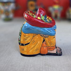 May include: A ceramic figurine of a person sitting with their head down, wearing a large red sombrero with floral accents, a blue shirt, and yellow pants. The figure has black outlines and painted details, showing a seated posture with legs tucked.