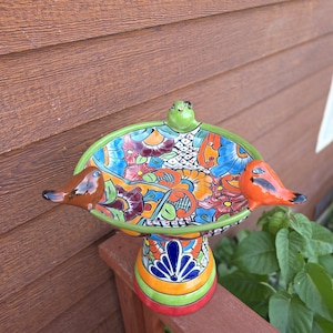 May include: A ceramic bird bath featuring a colorful hand-painted floral pattern in shades of blue, orange, red, and green. The bowl has three sculpted birds attached to the rim, colored in green, brown, and orange, and sits on a pedestal base.