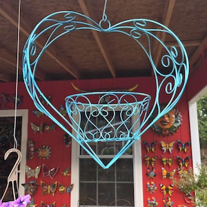 May include: A hanging, light blue metal planter shaped like a heart. The heart-shaped frame has decorative swirls and holds a basket-shaped planter. The planter is suspended from a hook, with a red wall and butterfly decorations in the background.
