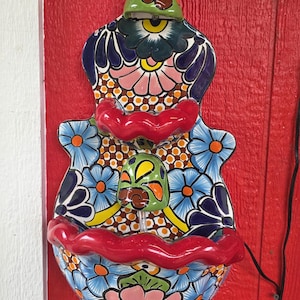 May include: A two-tiered ceramic wall fountain featuring a vibrant hand-painted floral pattern in shades of blue, pink, green, and orange. The piece has scalloped red edges on both basins and a central green spout, showcasing intricate Talavera-style details.
