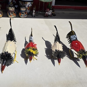 May include: Four metal rooster sculptures arranged in a row. Each figure features a long tail, a pointed beak, and a ruffled neck collar. The roosters are painted in various colors including black, white, red, and yellow, with distinct metallic textures.