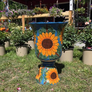 May include: A ceramic pedestal planter featuring a vibrant blue background adorned with hand-painted orange sunflowers and green leaves. The vessel has a wide, scalloped rim and a tapered base, showcasing a repeating floral motif throughout the exterior.