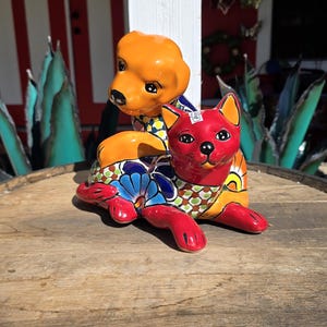 May include: A ceramic dog and cat figurine set. The dog is orange, and the cat is red, both with colorful floral patterns. The figurines are displayed on a wooden surface, with a red and white building in the background.