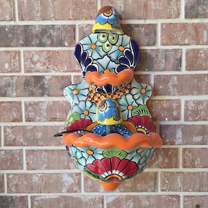 May include: A wall-mounted ceramic fountain featuring a vibrant, multi-colored floral pattern in shades of blue, green, red, and yellow. The piece has two scalloped, orange-rimmed basins and a central spout, with a black power cord visible.