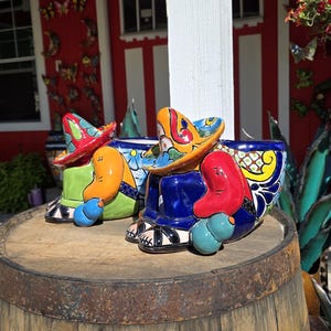May include: Colorful ceramic planters shaped like figures wearing sombreros. The planters are painted with bright colors including blue, orange, red, and green. They sit on a weathered wooden barrel, with a red building in the background.