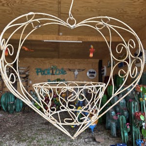 May include: Cream-colored, heart-shaped hanging planter made of wrought iron. The planter has a basket-like container at the bottom and decorative scrollwork throughout. A wire loop at the top allows for hanging. The background includes a shop with various decorative items.