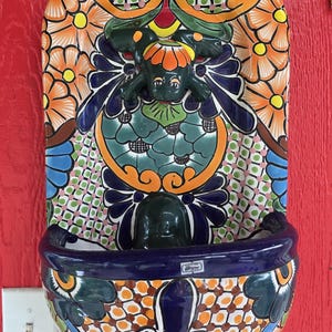 May include: A ceramic wall fountain featuring a hand-painted floral pattern in orange, blue, green, and white. The piece includes a central sculpted frog figure and a deep, rounded basin at the bottom with a small rectangular label on the front rim.