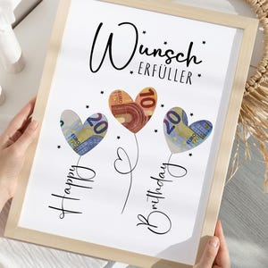 May include: A framed print with the words "Wunsch Erf&uuml;ller" and "Happy Birthday" in script. The artwork features three heart-shaped balloons made of Euro banknotes. The frame is light-colored.
