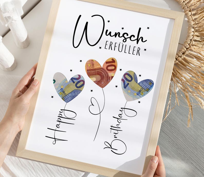 May include: A framed print with the words "Wunsch Erf&uuml;ller" and "Happy Birthday" in script. The artwork features three heart-shaped balloons made of Euro banknotes. The frame is light-colored.