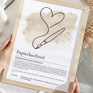 K&ouml;nnte beinhalten: Gerahmter Druck mit dem deutschen Wort "Papierhochzeit" und seiner Definition. Das Kunstwerk zeigt eine schwarze Strichzeichnung eines Stifts, der ein Herz zeichnet, mit einem beigen Aquarellhintergrund.