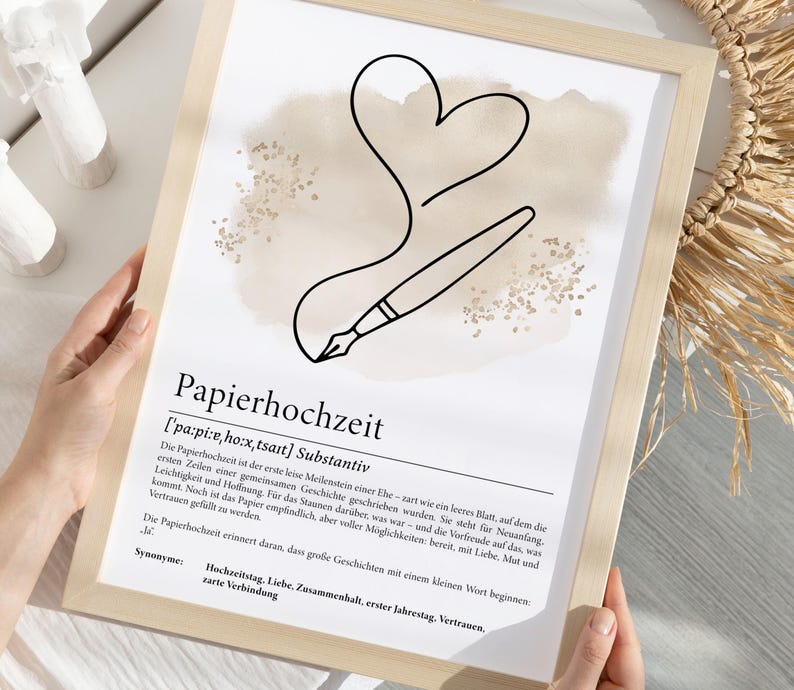K&ouml;nnte beinhalten: Gerahmter Druck mit dem deutschen Wort "Papierhochzeit" und seiner Definition. Das Kunstwerk zeigt eine schwarze Strichzeichnung eines Stifts, der ein Herz zeichnet, mit einem beigen Aquarellhintergrund.