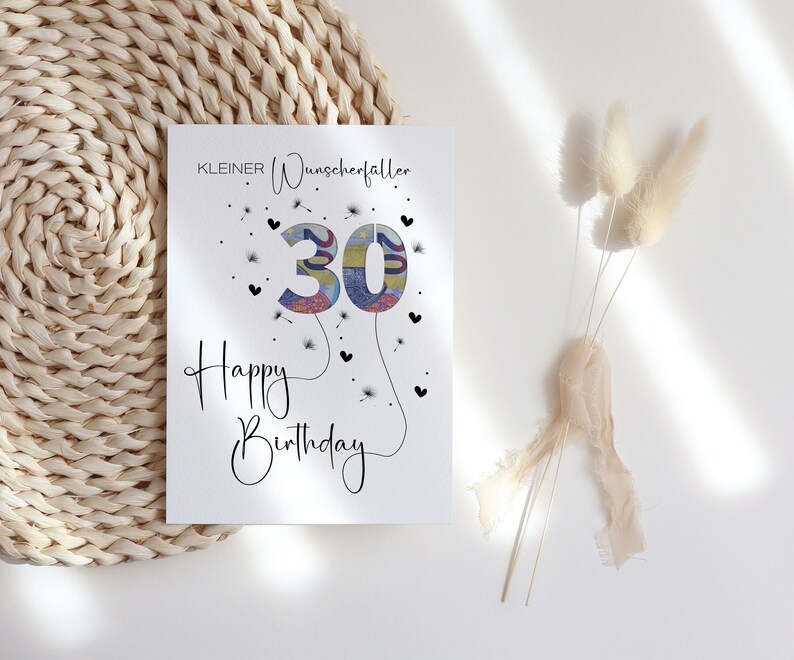 Peut inclure: Une carte blanche avec le texte "KLEINER Wunschertaller" et "Happy Birthday" avec le chiffre 30 fait d'argent au centre. Le chiffre 30 est entour&eacute; de petits c&oelig;urs noirs et de graines de pissenlit.