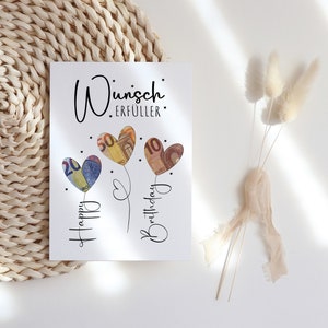 Bild mit der Aufschrift „Wunscherfüller“ in stilvoller, schwarzer Schrift. Darunter drei herzförmige Ballons, hinterlegt mit Euro-Geldscheinen (20, 50 und 10 Euro). Die Ballons sind mit feinen Linien verbunden, darunter steht „Happy Birthday“ in verschnörkelter Schrift. Kleine Sterne verzieren das Design auf weißem Hintergrund.