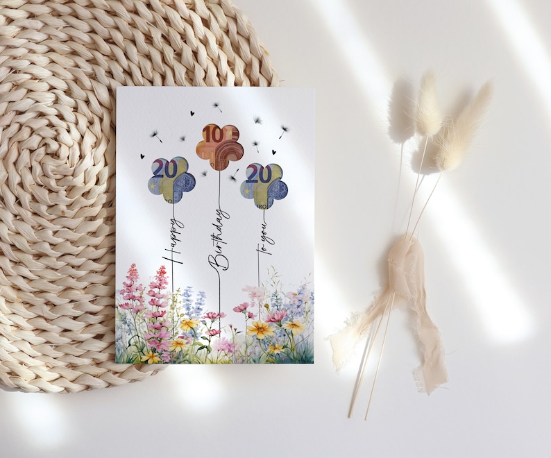 Blumiges Geldgeschenk zum Geburtstag als digitaler Sofort-Download. Drei Ballons  in Blumenform mit Euro-Scheinen hinterlegt steigen über einem bunten Wildblumenfeld in die Luft, begleitet von Pusteblumen und der Aufschrift „Happy Birthday to you“. Ein liebevolles, fröhliches Geburtstagsbild zum Ausdrucken – ideal als DIY-Geschenk in Form eines Bildes oder einer selbstgebastelten Karte für Blumenliebhaber.