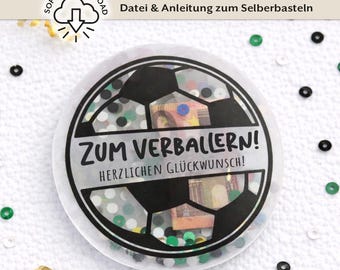 Puede incluir: Decoración redonda transparente con diseño de balón de fútbol, llena de confeti y divisas. El texto "ZUM VERBALLERN! HERZLICHEN GLÜCKWUNSCH!" está impreso. Fondo blanco con confeti disperso y elementos decorativos.