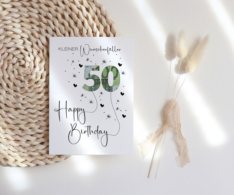 May include: A white card with black text that reads "KLEINER Wunscherfallen" and "Happy Birthday". The number 50 is made of a euro bill and surrounded by black hearts and dandelion seeds.