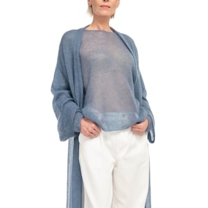 May include: A woman wearing a light blue, sheer, long-sleeved top and matching long cardigan. She is also wearing white pants. The clothing is made of a soft, flowing material. The outfit is a casual, yet elegant style.