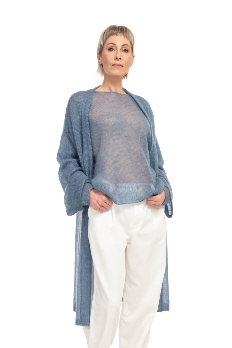 May include: A woman wearing a light blue, sheer, long-sleeved top and matching long cardigan. She is also wearing white pants. The clothing is made of a soft, flowing material. The outfit is a casual, yet elegant style.