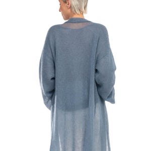 May include: A long, light blue cardigan with long sleeves. The cardigan is made of a sheer, open-knit fabric. The model is wearing the cardigan over a white top and pants. The cardigan falls to the mid-calf.