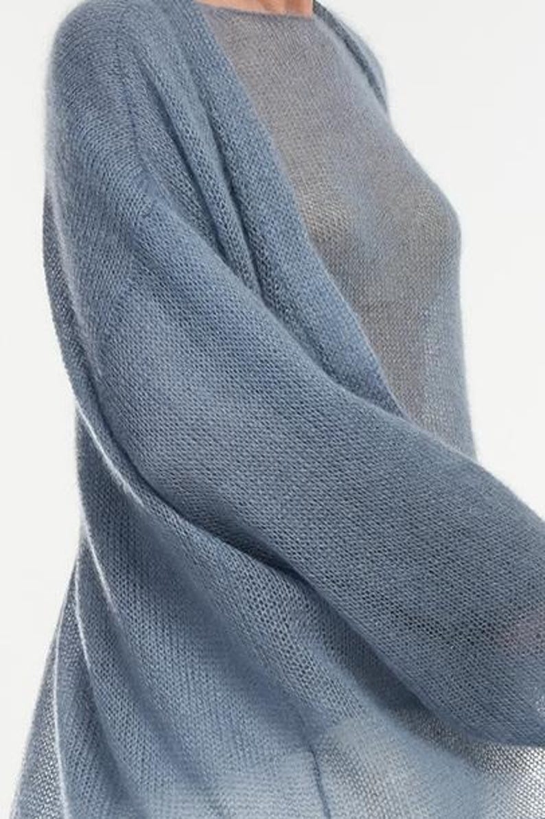 May include: A light blue knit cardigan with long sleeves. The cardigan is made of a soft, sheer material and drapes over a light gray top. The knitwear has a loose weave, creating a delicate texture. The overall style is casual and comfortable.