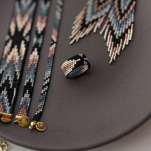 K&ouml;nnte beinhalten: Ein Set aus Perlen-Schmuck, darunter Armb&auml;nder, Ohrringe und ein Ring. Der Schmuck weist ein geometrisches Muster in Schwarz, Blau und Pfirsichfarben auf. Die Armb&auml;nder haben goldene Verschl&uuml;sse. Die Ohrringe haben ein Fransen-Design.