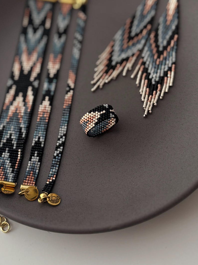 K&ouml;nnte beinhalten: Ein Set aus Perlen-Schmuck, darunter Armb&auml;nder, Ohrringe und ein Ring. Der Schmuck weist ein geometrisches Muster in Schwarz, Blau und Pfirsichfarben auf. Die Armb&auml;nder haben goldene Verschl&uuml;sse. Die Ohrringe haben ein Fransen-Design.