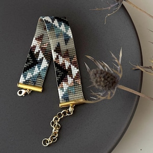 Könnte beinhalten: Ein Armband mit Perlenmuster in Grau-, Weiß-, Schwarz-, Braun- und Türkistönen. Das Armband hat goldfarbene Verschlüsse und eine Kettenverlängerung. Präsentiert auf einer dunkelgrauen Oberfläche. Ein stilvolles Accessoire.