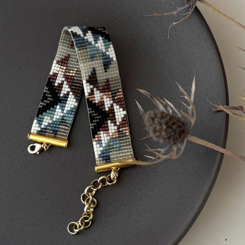 May include: A beaded bracelet with a geometric pattern in shades of gray, white, black, brown, and teal. The bracelet has gold-colored clasps and a chain extender. The bracelet is displayed on a dark gray surface.