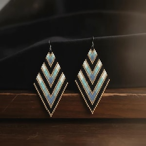 May include: A pair of diamond-shaped dangle earrings crafted from small beads in a chevron pattern. The design features alternating rows of black, light blue, and metallic gold beads, suspended from thin silver-toned metal earring hooks.