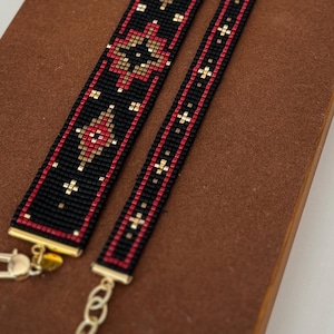 Puede incluir: Dos pulseras de cuentas con fondo negro y patrones geométricos en rojo y oro metálico. Las pulseras están terminadas con terminales de metal dorado, un cierre de mosquetón y una cadena de extensión ajustable para el ajuste.