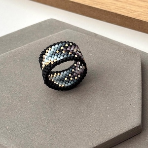 Könnte beinhalten: Ein Perlenring mit schwarzer Basis, verziert mit einem Muster aus blauen, goldenen und lila Perlen. Der Ring wird auf einer grauen Oberfläche mit einem Holzelement im Hintergrund präsentiert. Das Design des Rings ist aufwendig und detailliert.