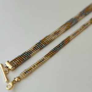 May include: A beaded necklace with a gold clasp and a geometric pattern in shades of brown, beige, and blue.