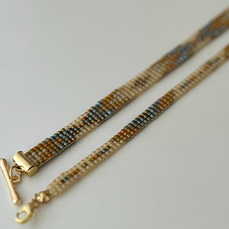 May include: A beaded necklace with a gold clasp and a geometric pattern in shades of brown, beige, and blue.