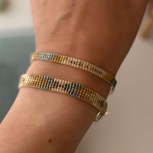 May include: Two beaded bracelets on a wrist. The bracelets are made with white, gold, and blue beads in a geometric pattern.