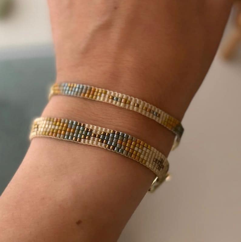 May include: Two beaded bracelets on a wrist. The bracelets are made with white, gold, and blue beads in a geometric pattern.