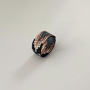 K&ouml;nnte beinhalten: Ein Perlenring mit einem geometrischen Muster in Schwarz, Grau, Kupfer und Silber. Der Ring besteht aus kleinen, gleichm&auml;&szlig;ig verteilten Perlen, wodurch eine strukturierte Oberfl&auml;che entsteht. Der Ring wird vor einem wei&szlig;en Hintergrund gezeigt.