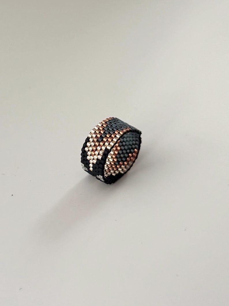 K&ouml;nnte beinhalten: Ein Perlenring mit einem geometrischen Muster in Schwarz, Grau, Kupfer und Silber. Der Ring besteht aus kleinen, gleichm&auml;&szlig;ig verteilten Perlen, wodurch eine strukturierte Oberfl&auml;che entsteht. Der Ring wird vor einem wei&szlig;en Hintergrund gezeigt.