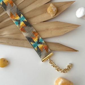 Può includere: Un braccialetto di perline con un motivo geometrico in tonalità di grigio, giallo, arancione e blu. Il braccialetto ha una chiusura dorata.