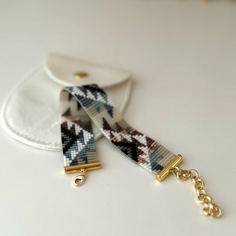 May include: A beaded bracelet with a geometric pattern in shades of blue, brown, and cream. The bracelet has gold-colored metal accents and a clasp. A small white pouch is in the background.