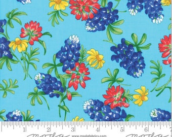 May include: Fabric with a repeating floral pattern featuring bluebonnets, red and yellow flowers, and green leaves on a light blue background. The fabric has a ruler along the bottom edge with the word "moda" printed on it.