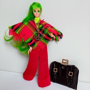 Op de afbeelding: Een pop met groen haar die een rood-groene poncho draagt met een vredespenning, een rode broek en een bruine leren koffer.