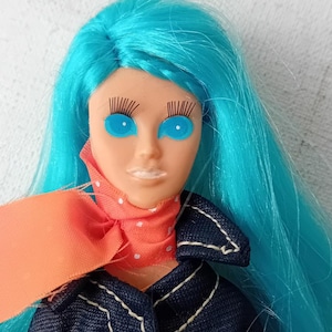 Op de afbeelding: Een vintage pop met lang, felblauw haar, die een spijkerjack met witte stiksels en een roze-witte stippen sjaal draagt. De pop heeft grote, blauwe ogen met lange wimpers.