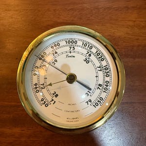 May include: A round brass barometer with a white face featuring black numerical scales for millibars, inches, and centimeters. The dial displays the text Boston, Chelsea Clock, and various pressure measurements with two black metal indicator needles.