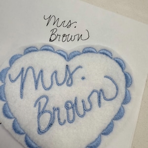 May include: A white heart-shaped patch with blue scalloped edging and embroidered text that reads "Mrs. Brown" in cursive. The patch is on a white surface, with the handwritten words "Mrs. Brown" above it.