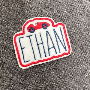 May include: White felt patch with red and blue stitching. The patch features a red toy car with a number "4" on the side and the name "ETHAN" in blue embroidery.