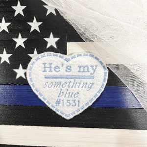 May include: A white felt heart with blue embroidery that reads "He's my something blue #1531". The heart is placed on a blue and white striped background with a white tulle overlay.