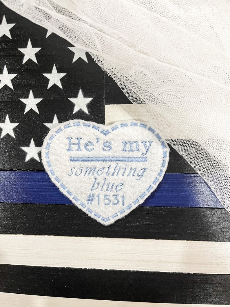 May include: A white felt heart with blue embroidery that reads "He's my something blue #1531". The heart is placed on a blue and white striped background with a white tulle overlay.