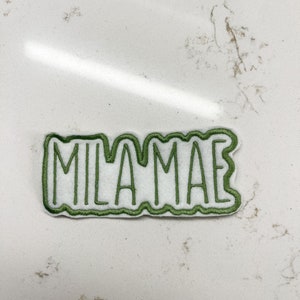 May include: Green embroidered patch with the word "MILAMAE" in a white background.